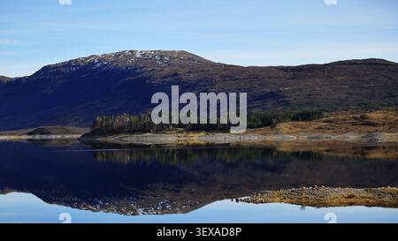 Loch Cluanie reflections in the Scottish Highlands, Scotland features low island, blue reflection, tree cluster, and Loch Cluanie across the scene. Stock Photo
