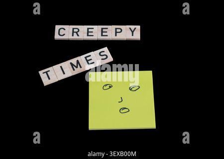 Black background holds word tiles: "CREEPY TIMES." Yellow sticky note has a simple, drawn face. Eyes and mouth suggest surprise or fear. The face star Stock Photo