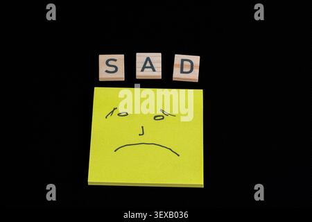 Black background shows simple sadness concept. Wooden blocks spell "SAD" above yellow sticky note. Hand-drawn face on note shows frowning expression Stock Photo
