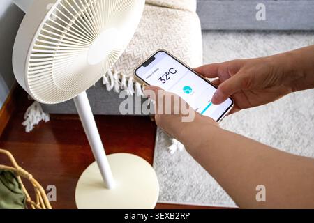 Hand holding a smartphone with a smart home application beside a pedestal fan inside a living room. Concept of wireless home automation, connected app Stock Photo
