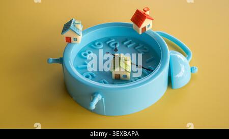 Three miniature toy houses arranged on top of a blue twin bell alarm clock over a seamless yellow background. Stock Photo