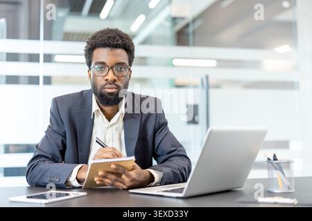 Professional man with a beard and glasses taking notes in a spiral notebook, sitting at a desk with a laptop and digital tablet, focusing on his work Stock Photo