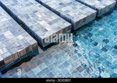 Luxury stone tiled lounge platform sit partially submerged channel in clear blue pool water and marble mosaic tiles covered floor Stock Photo