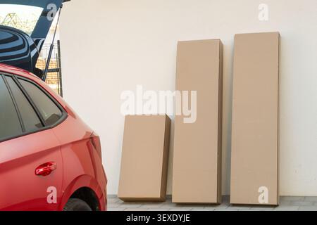 Stack of three cardboard boxes of varying sizes positioned beside a red car.  Stock Photo