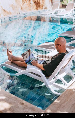 Top view of bald man freelancer CEO business owner working laptop on sun lounger by luxury hotel swimming pool. Digital nomad successfully managing co Stock Photo