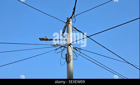 A cylindrical concrete utility post supports multiple overhead electrical cables, wire bundles, and a horizontal modern street lamp against a clear bl Stock Photo