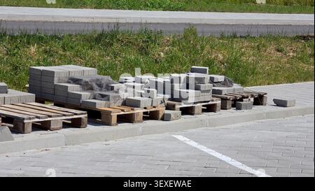 Rectangular gray concrete pavers and mesh netting sit stacked on wooden pallets next to a newly installed pedestrian walkway and grassy roadside curb Stock Photo