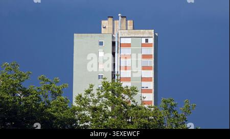 A light green high rise building with red brick window panels and roller shutters stands against a dark blue stormy sky background Stock Photo