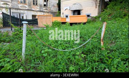 A low chain link barrier spans across lush green weeds and tall grass in front of an old neglected stone building courtyard with discarded orange Stock Photo