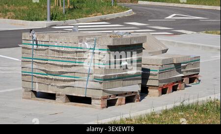 Three wooden shipping pallets hold neatly arranged piles of gray concrete masonry blocks wrapped in protective sheeting next to a fresh asphalt street Stock Photo