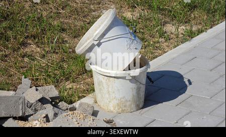 A pair of weathered white plastic pails smeared with dried mortar residue sit nested on a gray interlocking brick pedestrian path Stock Photo