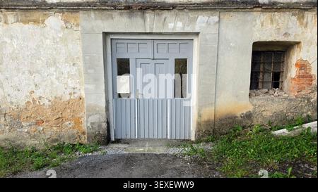 An old masonry building facade features a paneled wooden entrance door with a lower vertical slat barrier next to a rusty iron window Stock Photo