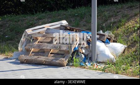 Several old wooden shipping pallets rest leaning against a heap of white filled woven bags and plastic wrapping debris along a gray paved pedestrian Stock Photo