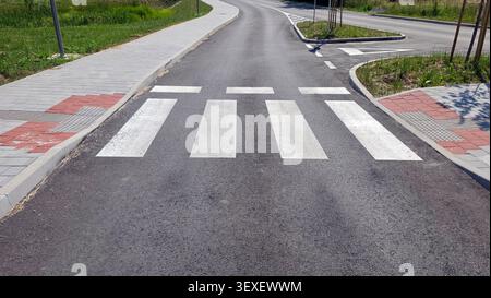 A pedestrian crosswalk with wide white stripes spans across a newly paved dark asphalt street connecting two gray and red stone block walkways with ta Stock Photo
