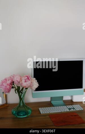 Elegant home workspace featuring pink peonies in a glass vase, desktop computer and clean wooden desk with copy space. Stock Photo