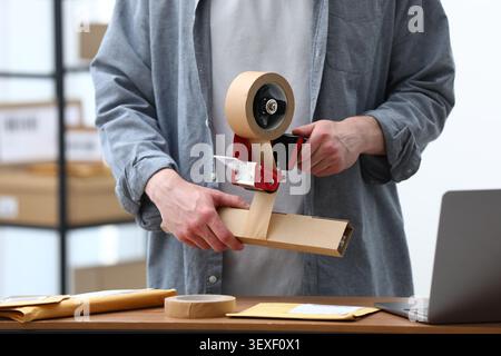 Man using dispenser with adhesive tape to seal cardboard box before sending at table in office, closeup Stock Photo