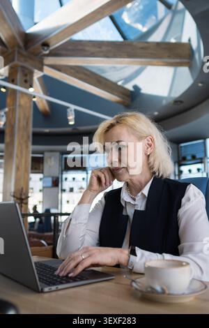 Elderly Businesswoman Reviewing Documents Near Laptop, Urban Cafe View In Background, Focused Planning Posture, Gentle Natural Light, Hands On Stock Photo