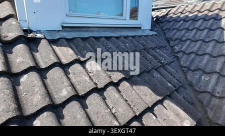 A view of dirty and aged grey concrete roof tiles surrounding a white uPVC dormer window. Stock Photo
