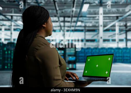 Data center engineer uses a green screen laptop in a data center for failure detection, screen replacement, and server workflow monitoring. Stock Photo