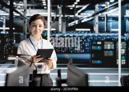Smiling data center engineer uses a tablet to review server infrastructure, diagnostics and AI technology in a data center. Stock Photo