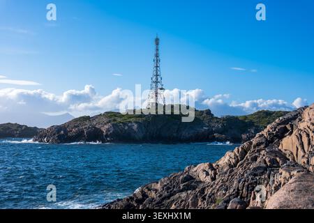 Tall red and white communication tower stands on a rocky, vegetation-covered island in Lofoten. Dramatic coastal landscape with deep blue Stock Photo