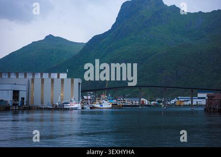 Scenic Lofoten fishing harbor with moored boats, industrial buildings, and dramatic green mountains backdrop under cloudy sky. Arctic Stock Photo
