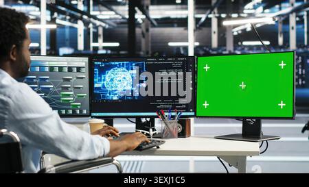 Data center specialist in wheelchair uses a green screen mockup computer in a server room for AI workflow and screen replacement. Stock Photo
