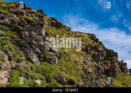 Steep rocky mountainside blanketed in vibrant green moss and lichen in Lofoten, Norway. Dramatic natural landscape with blue sky showcasing Stock Photo