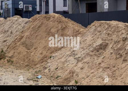 Workers are using sand piles for construction on a site surrounded by buildings during the day. Dust is visible in the air. Stock Photo