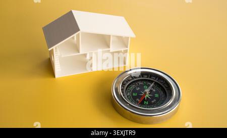 High angle view of a white model house frame section placed next to a metal magnetic navigation compass on a plain yellow surface. Stock Photo