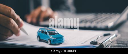 Person signing buying car contract for vehicle purchase and insurance, close up of hand with pen on document, small blue toy car on clipboard, laptop Stock Photo