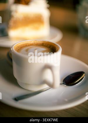 Steaming white coffee cup with smooth foam sits on a saucer. Silver spoon rests nearby. Blurred cake pieces appear in the background. Warm light creat Stock Photo