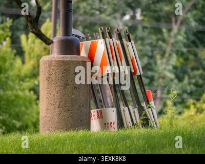 Folded orange and white striped traffic construction barricades stacked against a concrete light pole base on a green grass hill Stock Photo