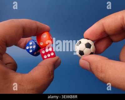Two hands compare a tiny soccer ball against three colorful rolling dice on a dark blue background. Stock Photo