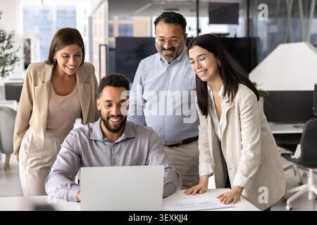 Four positive diverse teammates gathered around laptop in modern office Stock Photo