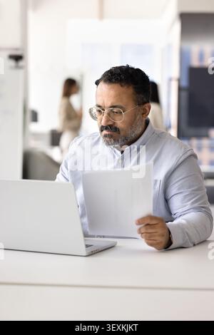 Businessman reviewing printed financial documents seated at workplace with laptop Stock Photo