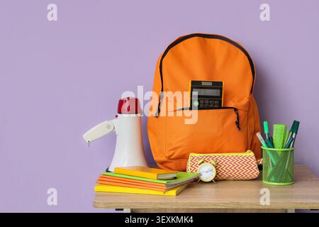 Orange backpack with school supplies on wooden table against purple wall Stock Photo