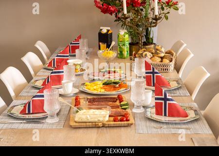 Long wooden table set for a festive Norwegian meal. Red and blue flags adorn each place setting. Fresh bread, cheese, and colorful vegetables are disp Stock Photo