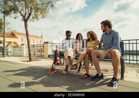 Multicultural group of young friends sitting together on a bench and talking outdoors in a sunny Mediterranean city during summer. Stock Photo