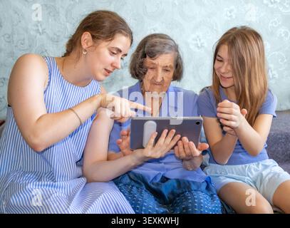 Family generations bonding as granddaughter and daughter teach grandmother to use a tablet at home Stock Photo