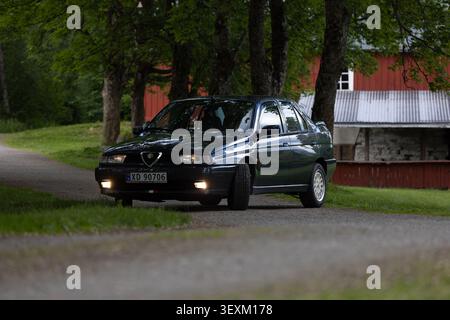 Steinkjer, Norway - June 15th 2024 - Dark Alfa Romeo 155 glides along quiet country road. Trees frame the scene with lush greenery. Red barn stands so Stock Photo
