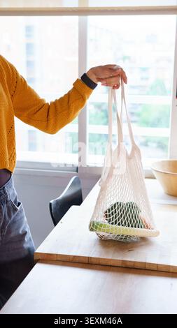 Person holding a reusable mesh bag filled with fresh healthy vegetables in a kitchen Stock Photo
