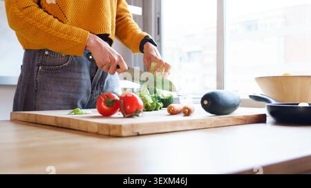 Woman's hands chopping fresh vegetables on a wooden cutting board in a brightly lit kitchen Stock Photo