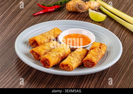 Golden fried spring rolls arranged on light gray plate with orange dipping sauce. Red chilies, lime wedge, lemongrass, galangal in background. Warm li Stock Photo
