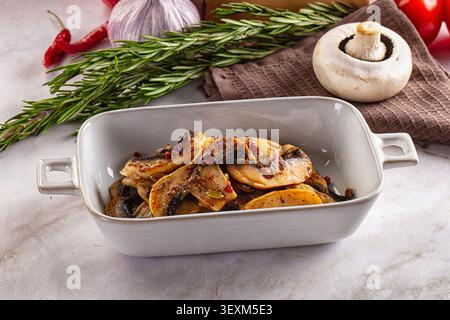 Sliced roasted mushrooms in white ceramic dish with rosemary sprigs garlic chili pepper on marble surface warm lighting earthy tones rustic food styli Stock Photo