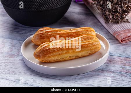 Golden baked eclairs on white ceramic plate. Rustic wood surface with muted blue gray tones. Soft natural lighting highlights pastry texture. Dried la Stock Photo