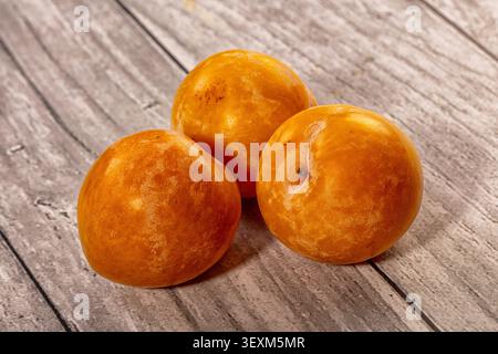 Three glossy yellow plums arranged on weathered gray wood. Warm natural lighting highlights fruit texture rich orange tones Stock Photo