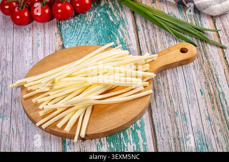 Cheese sticks arranged on round wooden cutting board. Red cherry tomatoes green onions in background. Rustic weathered wood surface with teal gray ton Stock Photo