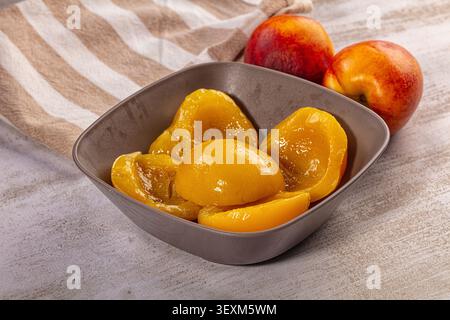 Fresh sliced peaches glisten in gray ceramic bowl beside whole ripe peaches on textured surface with beige white striped cloth. Warm golden yellow red Stock Photo