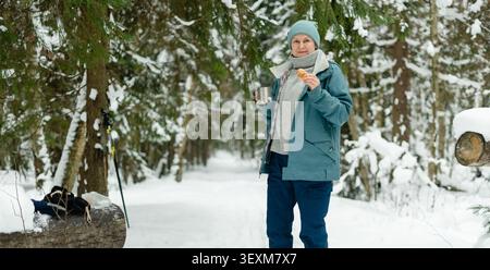 Middle-aged lady having a coffee break on a panoramic winter forest trail. Forty-something woman holding a steaming cup near a log on a snowy path. Horizontal banner with expansive cold woodland view Stock Photo